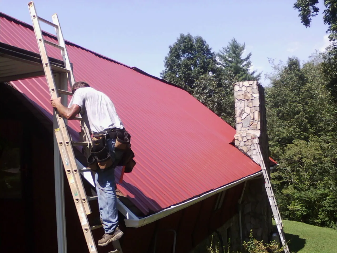Roofer on a ladder beside a red metal roof and stone chimney — Soffit & Fascia Repair by Roofing Brothers in Blakely